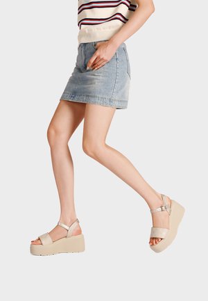 Donna che indossa sandali con zeppa beige, mini gonna in denim chiaro e top senza maniche a righe, cammina su uno sfondo chiaro uniforme.