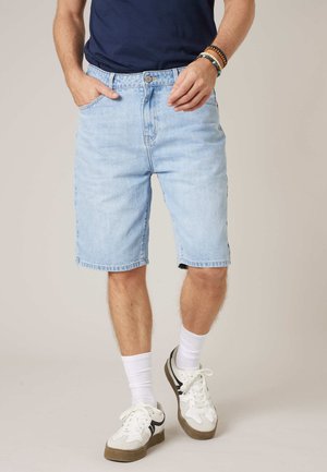 Man draagt lichtblauwe denim shorts, witte sneakers, witte sokken, een marineblauw shirt en kralenarmbanden, staand met handen dicht bij de zakken.