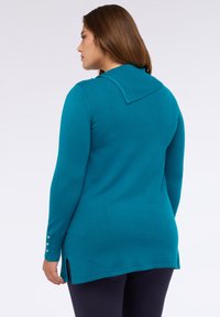 Teal tröja med långa ärmar och ribbat krage, med sidofickor och gyllene knappr detaljer vid ärmsluten. Slät yta.