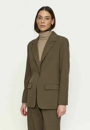 Femme aux cheveux bruns lisses porte un costume vert olive et un col roulé beige, debout sur un fond blanc uni, face vers l'avant.