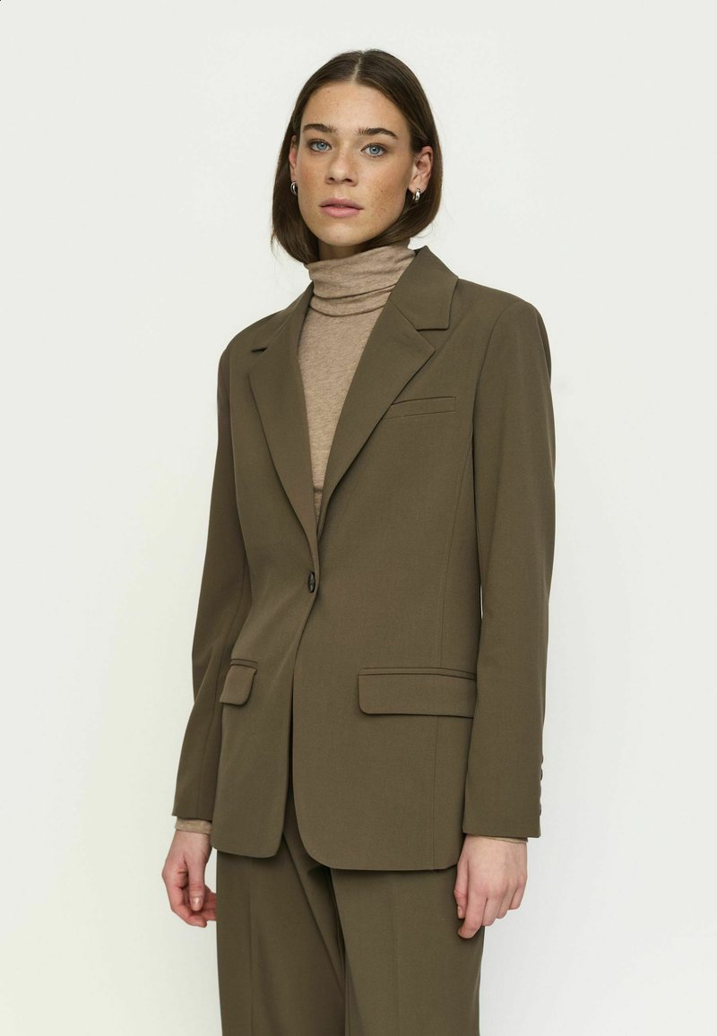 Femme aux cheveux bruns lisses porte un costume vert olive et un col roulé beige, debout sur un fond blanc uni, face vers l'avant.