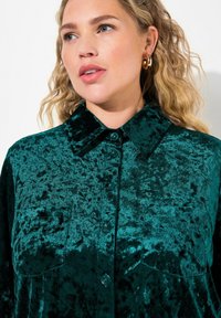 Camisa de terciopelo verde con cuello, que presenta un cierre de botones en el frente y dos bolsillos en el pecho. Textura suave y un brillo sutil evidente en la tela.