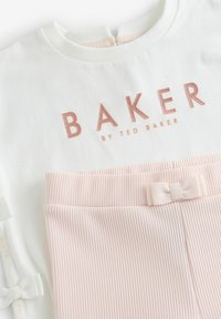Balta kokvilnas blūze ar "BAKER" rozā zelta burtiem, apvienota ar rozām ribotām biksēm, kurām jostas vietā ir neliels zieds.