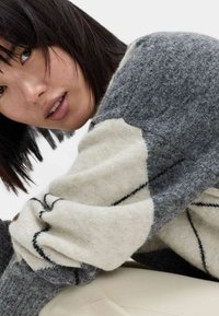 Fleckiger Pullover mit einem grau- und cremefarbigen Blockdesign, das über eine weiche Textur, schwarze Nähakzente und einen runden Halsausschnitt verfügt.