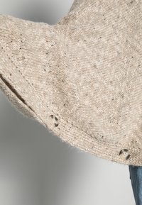 Beiger, gestrickter Pullover mit strukturiertem Stoff, versehen mit einer Knopfdetail am Saum und einem diagonalen Strickmuster. Die Kanten haben eine leichte Kräuselung.