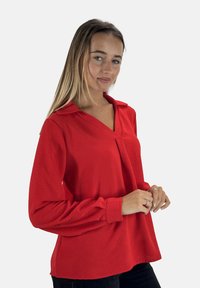 Blusa rossa a maniche lunghe con colletto a V e parte anteriore plissettata. Tessuto morbido e texture. Maniche con polsini e vestibilità comoda. Sono indossati pantaloni neri.