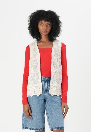 Cream-colored lace vest with intricate patterns, worn over a red long-sleeve shirt, paired with blue denim shorts. Textured and layered design.