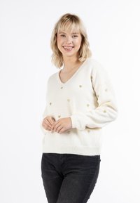 Crème V-hals trui van zachte stof, met een patroon van kleine gouden hartjes erdoorheen. Geribbelde manchetten en zoom. Gemaakt met donkere jeans.
