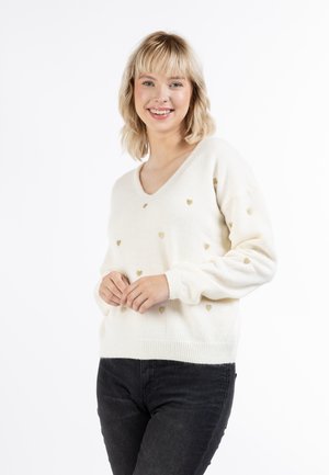 Pulover crem cu decolteu în V, realizat dintr-un material moale, având un model cu mici inimi aurii pe întreaga suprafață. Manșete și tiv cu nervuri. Purtați cu blugi negri.