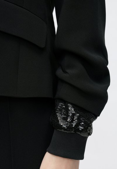 Must kohandatud jakk struktuurse disainiga, millel on tekstuuriga litritega must mansett ja soonikkude detail randmel.