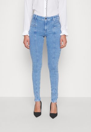 Slim-fit lichtblauwe spijkerbroek met verticale naden, gecombineerd met een witte blouse met geschulpte randen en zwarte puntige hakken.