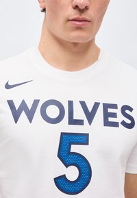 Tuva personāls ar baltu sporta krekliņu, uz kura priekšpuses ir uzraksts "WOLVES" un liels zils skaitlis 5.