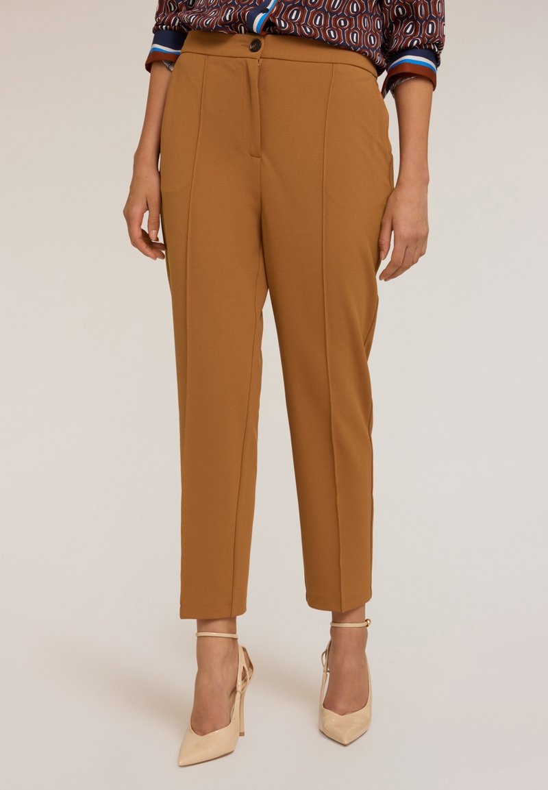 Femme portant un pantalon chevillard camel sur mesure avec pli devant, associé à des chaussures à talons beiges et une chemise à motifs.