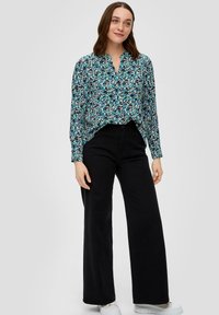 Langarmshirt in blauw met een bloemenpatroon en marineblauwe, groene en zwarte accenten; gecombineerd met zwarte wijde broek en witte sneakers.