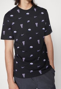 Musta Nike t-paita, jossa on violetti "Just Do It." -teksti, Nike-logoja ja koripalloteemoja, jota henkilö pitää kädet taskuissa.