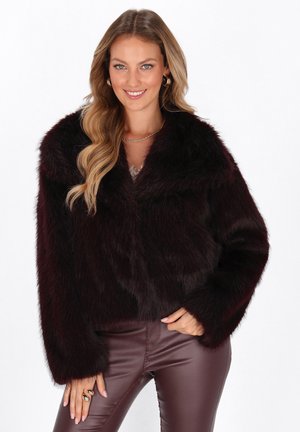 Vrouw in een donkerbordeaux nepleren bontjack en bijpassende leren broek, glimlachend met haar hand op haar heup tegen een witte achtergrond.