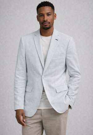 Homme portant un blazer gris clair sur un t-shirt blanc et un pantalon beige, debout avec une main dans la poche contre un fond uni.