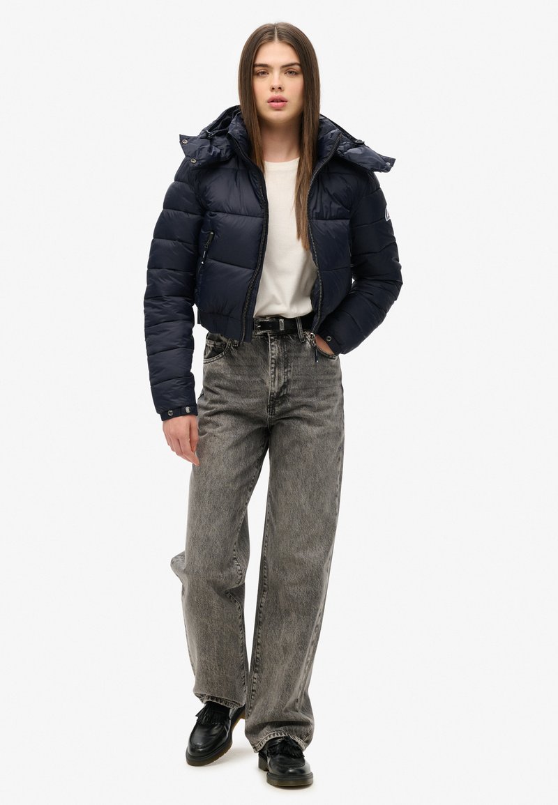 Marineblauwe pufferjack met capuchon, ritssluiting en geborduurde textuur. Gecombineerd met hoog opgesneden grijze jeans en zwarte schoenen. Eenvoudig wit shirt eronder.