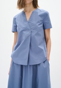 Blusa azzurra chiara con maniche corte, scollatura a V e dettaglio con pieghe frontali. Realizzata in tessuto morbido, abbinata a una gonna coordinata.