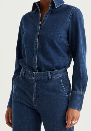 Kvinde iført en mørkeblå denim skjorte med lange ærmer og knapper, stukket ned i matchende højtaljede denim bukser med bæltestropper.