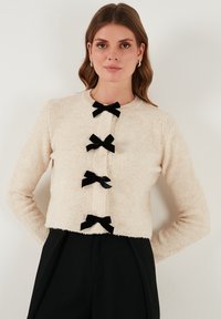 Maglione corto beige chiaro realizzato in morbido tessuto testurizzato, con quattro fiocchi neri in velluto come chiusure e un collo rotondo.