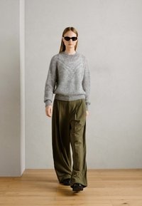 Grauer gestrickter Pullover mit Rippstruktur, kombiniert mit lockeren olivgrünen Hosen. Das Model trägt schwarze Sonnenbrillen und schwarze Schuhe.