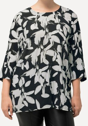 Femme portant un chemisier noir ample avec un motif floral abstrait blanc et des manches trois-quarts, associé à un pantalon noir.