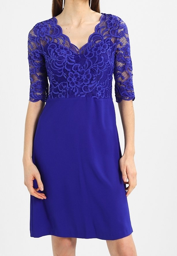 Femme portant une robe bleu roi longueur genou avec un corsage en dentelle florale et des manches mi-longues, encolure en V, debout sur un fond uni.