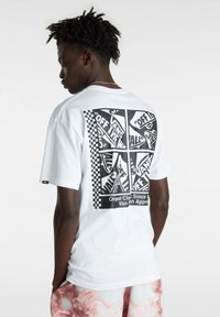 Camiseta de algodón blanca con un gran gráfico negro en la parte posterior que presenta logotipos y un patrón de tablero de ajedrez. Mangas cortas, ajuste relajado.