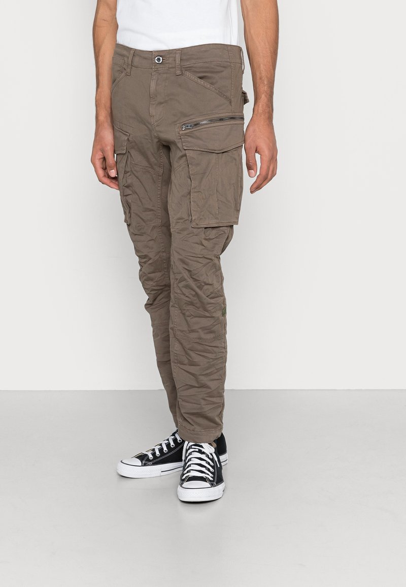 Homme portant un pantalon cargo marron avec plusieurs poches et des baskets montantes noires, debout devant un fond clair uni.