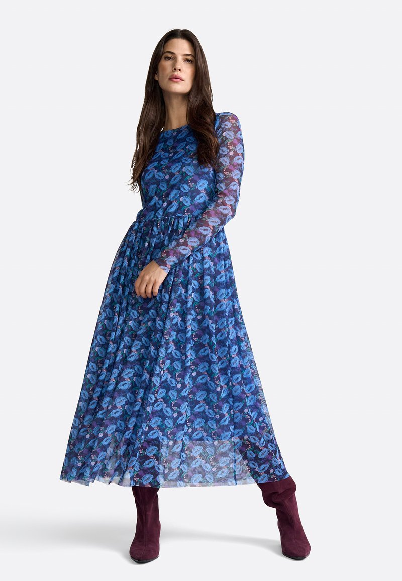 Vestido maxi azul de flores con mangas largas, superposición transparente, cintura fruncida y botas moradas hasta el tobillo. El patrón presenta diferentes tonos de azul y verde.