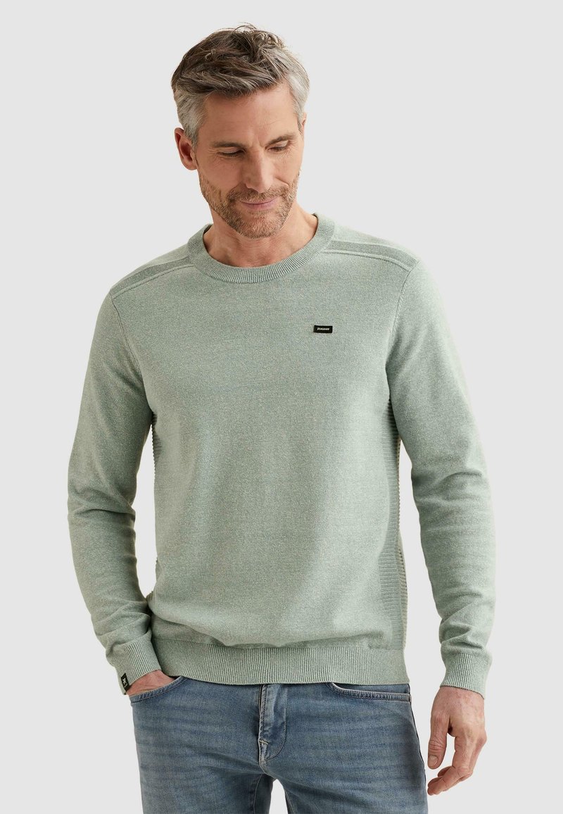 Homme aux cheveux gris portant un pull vert clair à col rond et un jean bleu, debout avec une main dans la poche, regardant vers le bas.