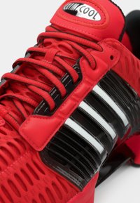 Red athletic shoe with mesh upper, black synthetic accents, and white-striped detailing. Textured laces and logo on the tongue.