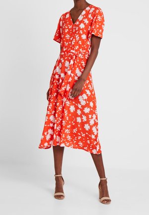 Mujer con vestido rojo hasta la rodilla con estampado floral blanco, mangas cortas, escote en V y cinturón en la cintura, combinado con sandalias de tacón alto beige.