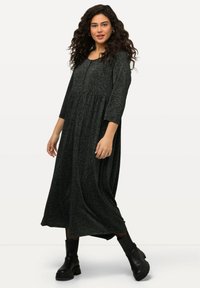Robe maxi noire à manches longues en tissu texturé, avec un bodice ajusté et une jupe évasée. Portée avec des bottes noires à chevilles.