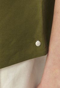 Cămașă verde măslinie dintr-un material texturat, având un etichetă metalică pe tiv. Cusături în contrast pe margine adaugă detalii.