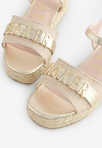 Gouden espadrillesandalen met brede gevlochten banden met "BAKER" in metallic letters. Gladde, glanzende zolen en een ingewikkeld gevlochten plateau.