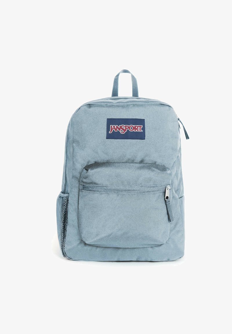 Jasnoniebieski plecak Jansport o prostokątnym kształcie, wykonany z materiału, z przednią kieszenią na zamek i naszywką z logo. Boczne siatkowe kieszenie na butelki z wodą.
