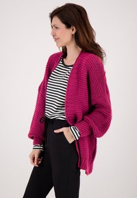 Fuchsia gebreide cardigan met een loose fit, geribde manchetten en open voorkant, gedragen over een zwart-wit gestreept shirt en zwarte broek.