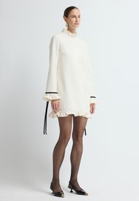 Abito color crema con colletto e maniche a volant, dettagli neri ai polsini e orlo a volant. Indossato con scarpe nere a punta.