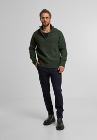 Donkergroene, gebreide pullover met een ritskraag, zwarte broek en zwarte laarzen. De pullover heeft een relaxte pasvorm en geribde manchetten.