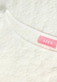 Witte kanten top met een gegolfde halslijn en een zachte textuur. Bevat een roze "JJXX" label nabij de kraag.