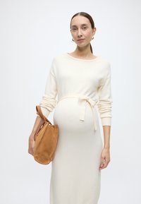 Κρεμ μάλλινο φόρεμα με μακριά μανίκια, ζώνη στη μέση. Κρατώντας μια ταμπά σουέντ τσάντα με κυρτό σχήμα. Ομαλή υφή σε όλο το φόρεμα.