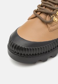 Botte de randonnée en cuir beige avec embout en caoutchouc noir, semelle crantée épaisse, et lacets marron et noir à motifs passant à travers des œillets dorés.