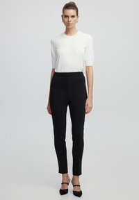 Pantaloni neri aderenti con una texture liscia, abbinati a una maglietta bianca a maniche corte. Il modello indossa tacchi neri con cinturini.