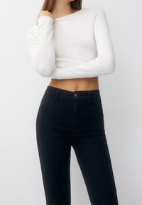 Mujer con una blusa larga blanca sin hombros y pantalones negros de cintura alta, de pie frente a un fondo blanco liso.