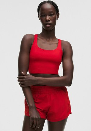 Kvinde med flettet hår iført rød ærmeløs crop top og matchende røde shorts, stående med den ene arm krydset over kroppen.