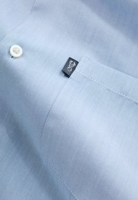 Camicia azzurro chiaro con bottoni, realizzata in tessuto con trama strutturata, presenta un bottone bianco e un'etichetta nera con logo argentato sulla tasca.