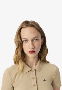 Polo shirt beige a coste con colletto, maniche corte e logo verde Lacoste. Spacchetto a cinque bottoni sul davanti.