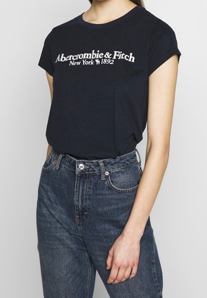 Vrouw die een zwart Abercrombie & Fitch T-shirt draagt, dat in een hoge blauwe spijkerbroek is gestopt, staand tegen een effen achtergrond.